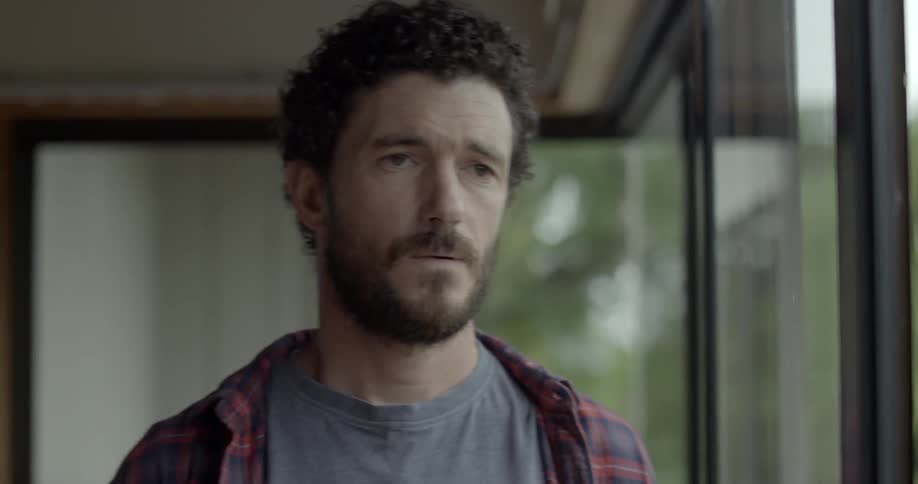 A bearded man in a plaid shirt looks out of a window and walks away