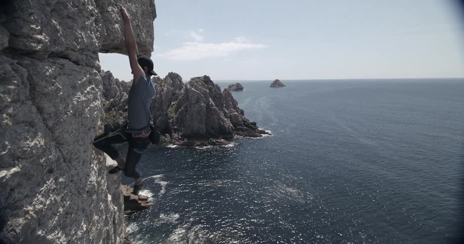 Young adult male hanging on to the side of a rock cliff with feet dangling above the ocean water on a bright day