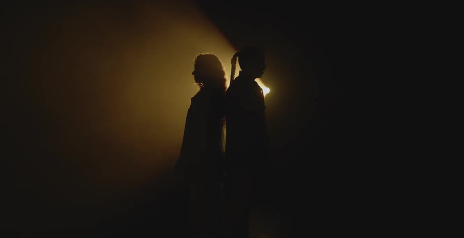 Two women in silhouette standing back to back backlit by a yellow light