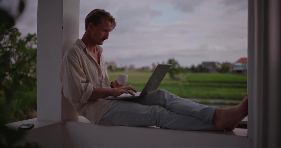 Comfortably dressed barefoot man sits on the ledge of a wall on an outdoor porch of a countryside villa at dusk tying on a laptop and sipping coffee
