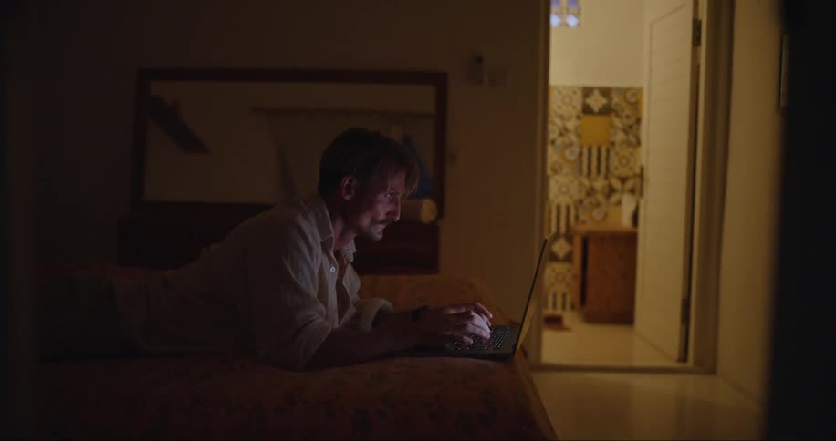 A man is lying across his bed typing on a laptop computer