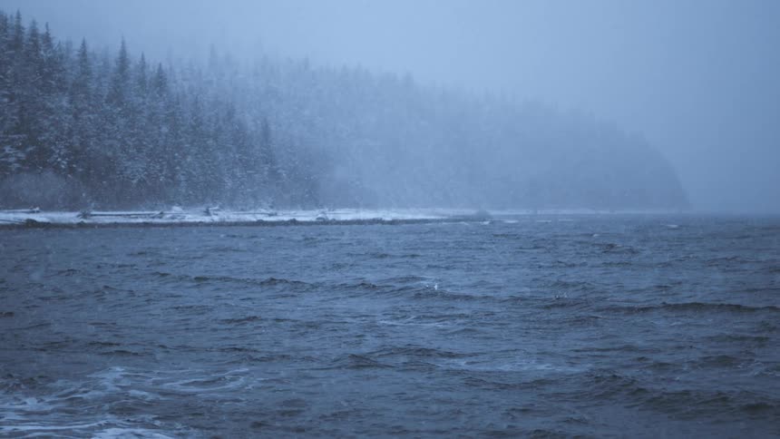 Snow falls over the ocean next to the mountains