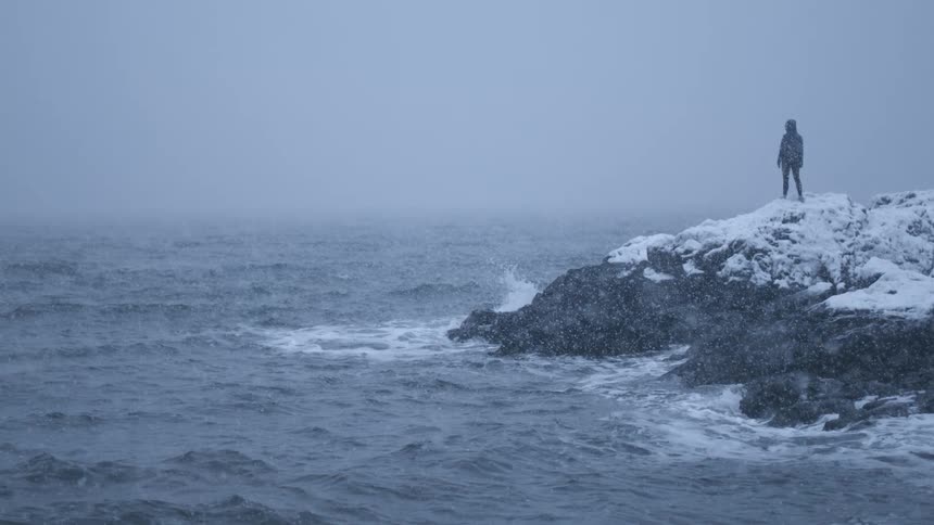 A female person stands on snow covered rocks beside a rough sea as snow falls