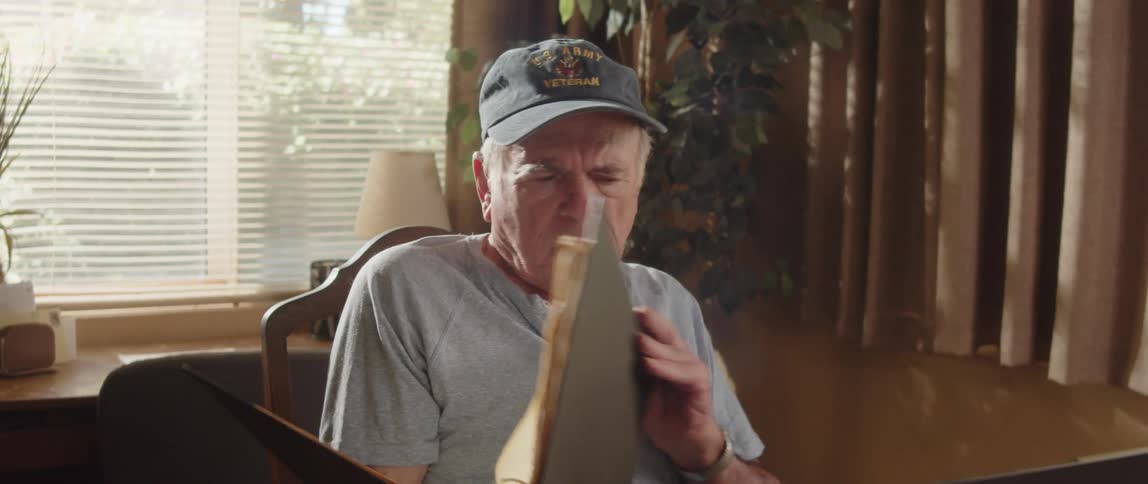 An elderly man is sitting at a table looking through a notebook and then at a laptop computer