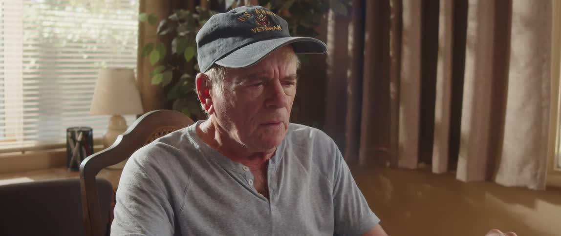 An older man sits indoors while scratching his face and coughing