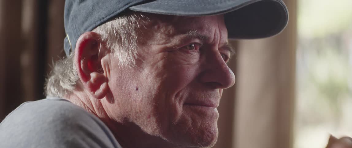 An older man puts on a us veterans ball cap and smiles while sitting indoors
