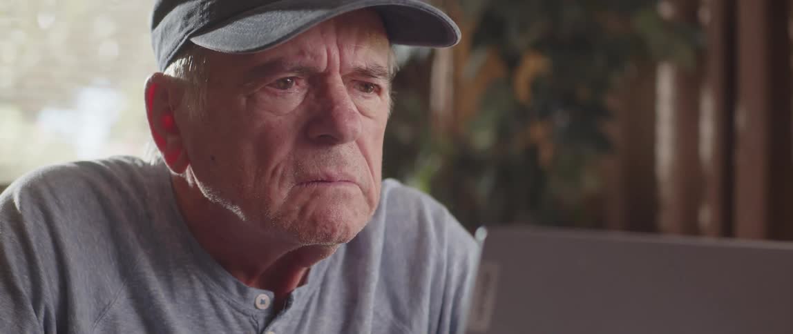 Elderly man sits and frowns after comparing spreadsheet document to laptop