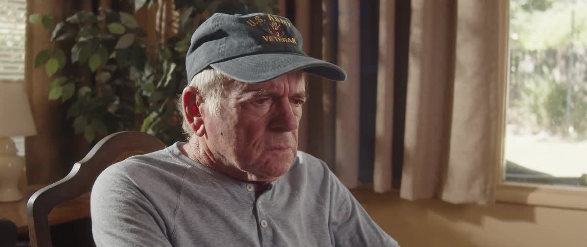 An elderly man is sitting near a window thinking as he puts on his cap