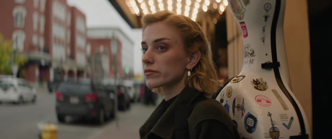 Female musician carrying cello case stands under theater lights outside looking at road