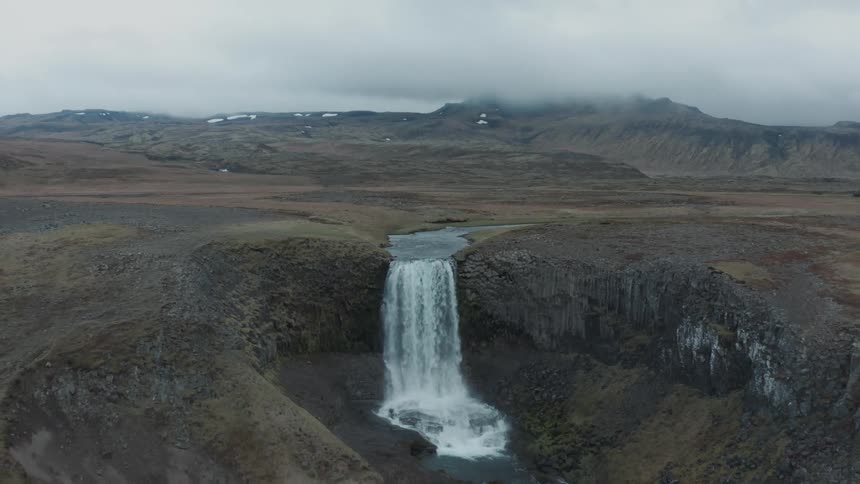 Waterfall emptying into a crater surrounded by barren volcanic highlands with cloud covered mountain in the background