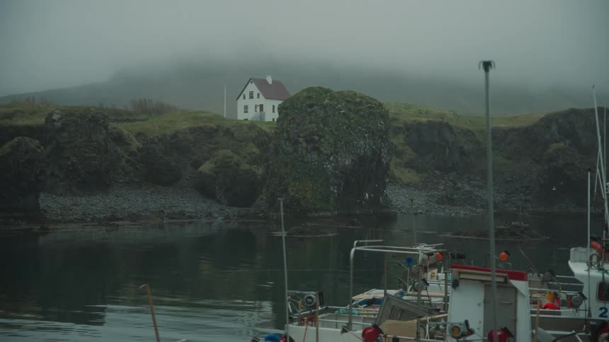 Boats docked near a shoreline with many rocks and a lone house in the fog