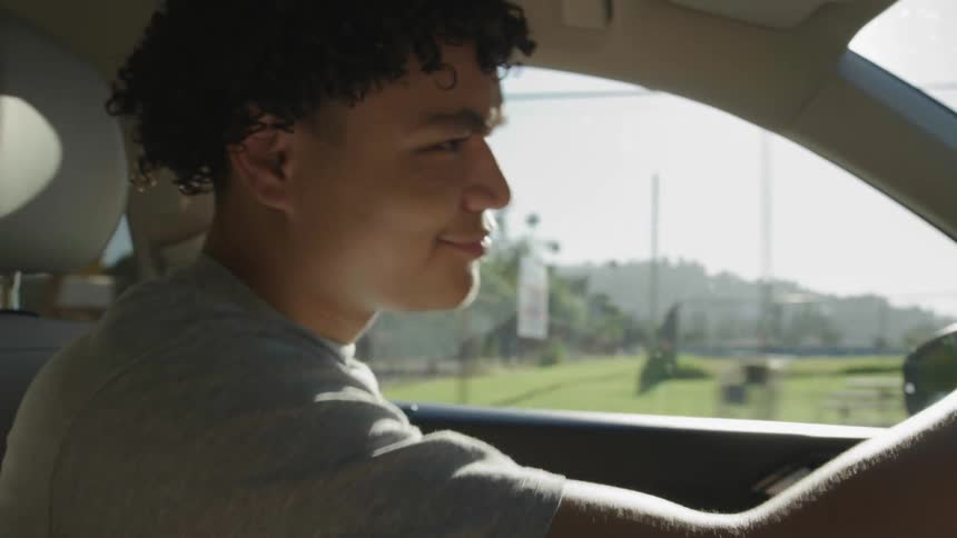Teenager with curly hair and a gray t shirt drives a car listening to music