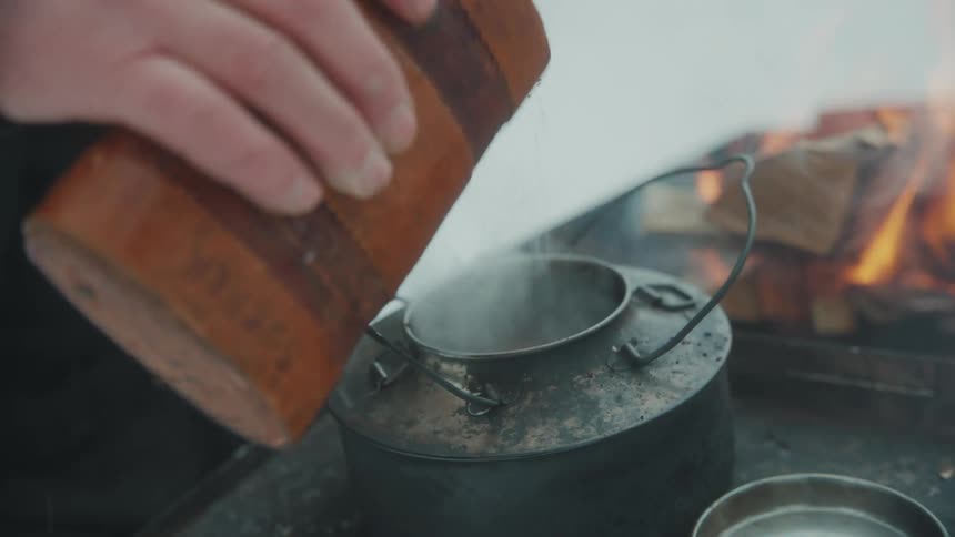A person pouring ground coffee into a pot over a wood stove