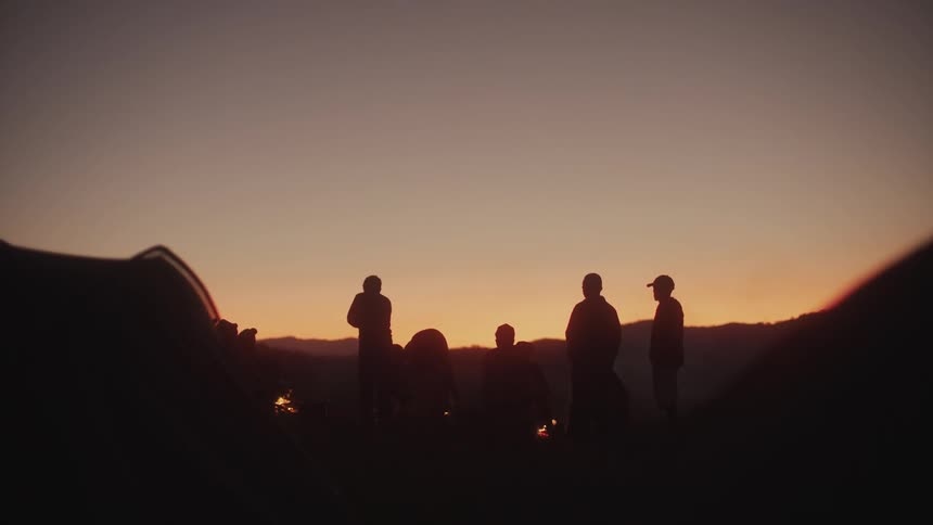 A group of silhouettes of people near bonfires and the sky is dawning