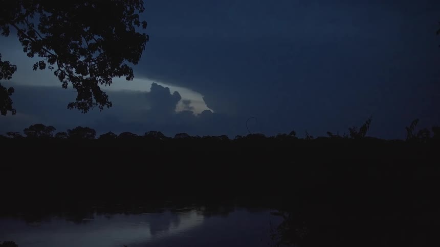 A storm in the sky with some lightning between clouds and there is a lake nearby