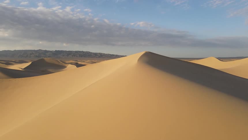 Low aerial view around of a pointed sand dune hill surrounded by sandy hills on a bright day orbiting from sun at the rear to sun straight ahead