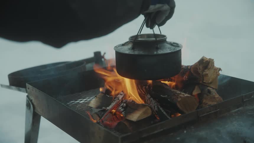 A mans arm is shown as he warms a kettle on a fire outside in the snow