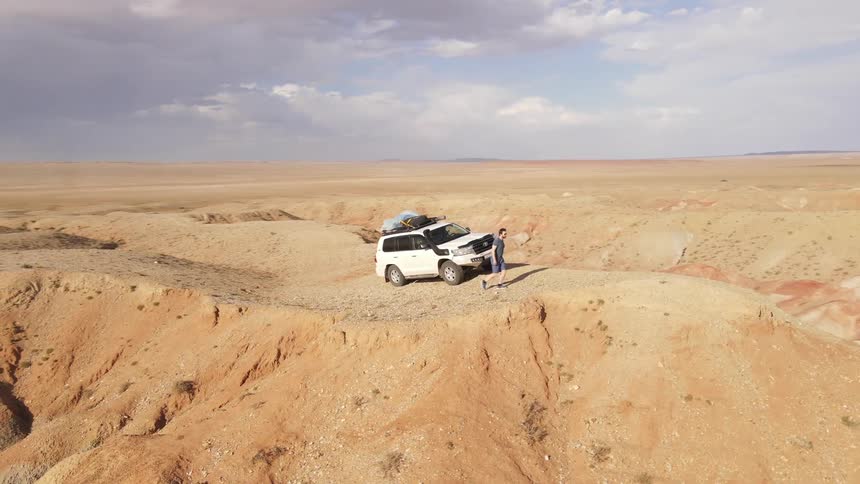 A young man steps out of a white suv that is parked on a plateau in the desert