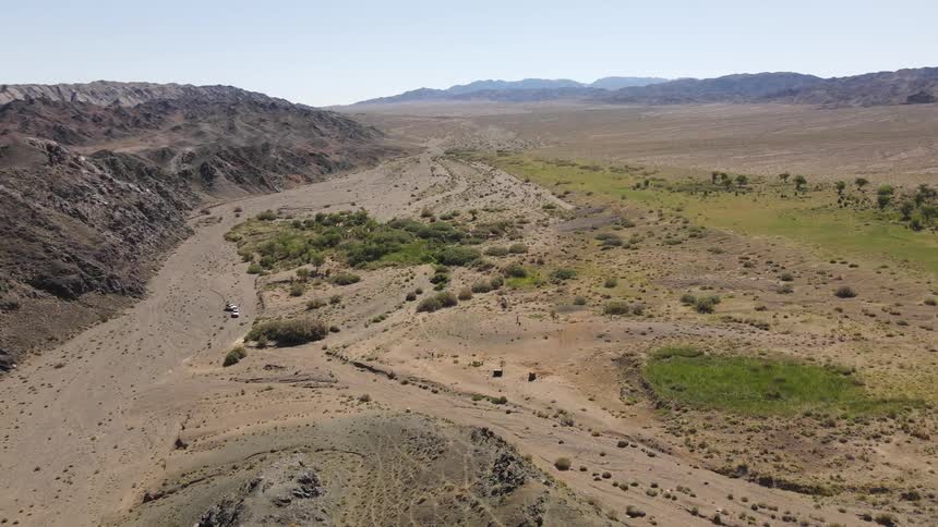 Aerial of vehicles parked on a trail around light vegetation area in a desert valley surrounded by hills on a bright day