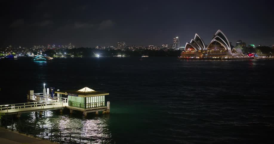 Water near pier reflects pier lights across harbor from the sydney opera house at night
