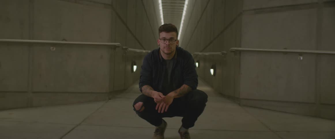 A squatting man snaps and taps his foot to a rhythm in a concrete hall