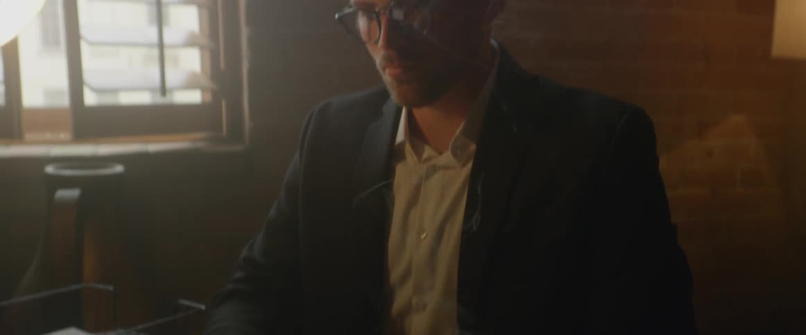 A man types at a typewriter as a cigarette burns on the desktop