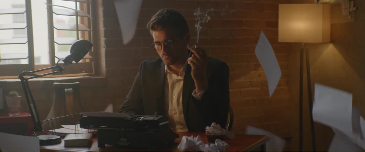 Paper falls around a man sitting in front of a typewriter smoking a cigarette