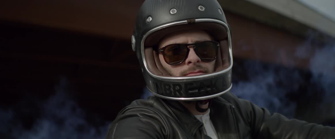 A man wears sunglasses and a motorcycle helmet as smoke rises behind him