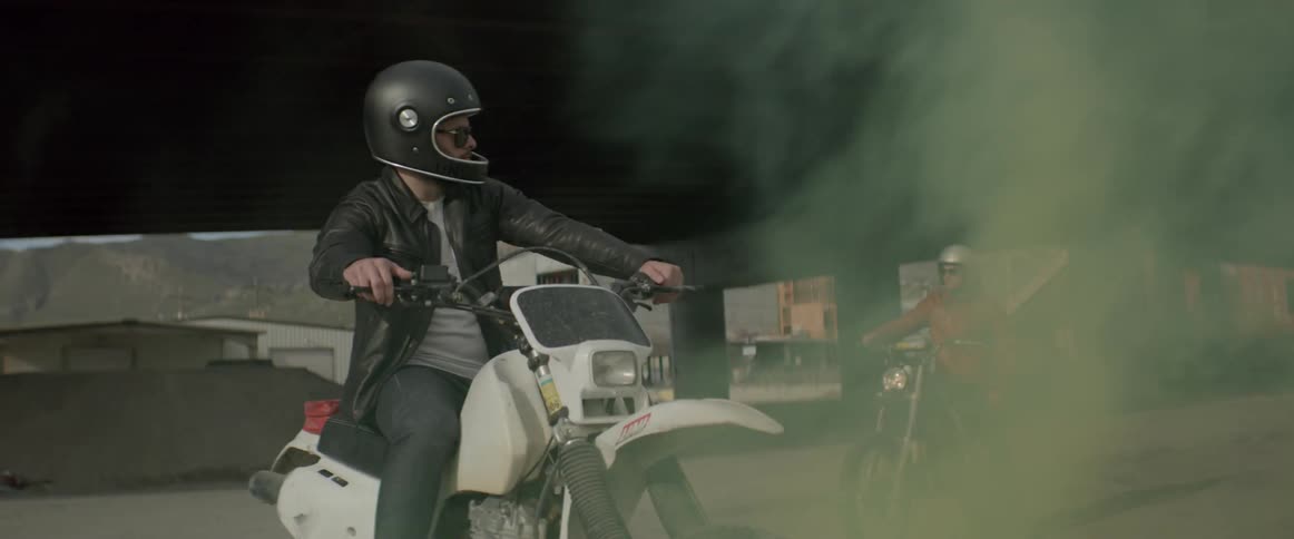 Two men sit on motorcycles while smoke blows in front of them