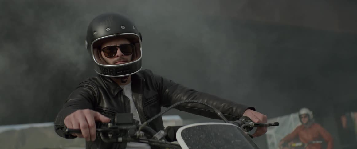 A man sitting on a motorcycle wearing a helmet turns and looks toward the camera