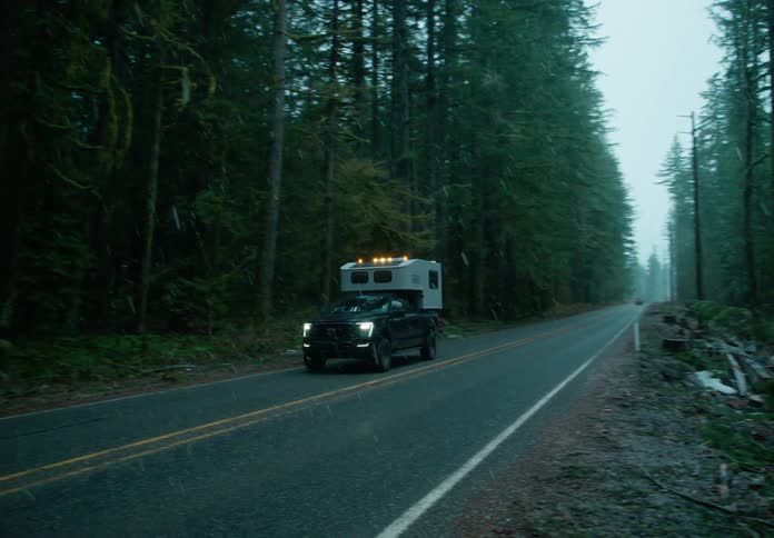 A pickup truck with a camper shell is driving on a road surrounded by dense forest