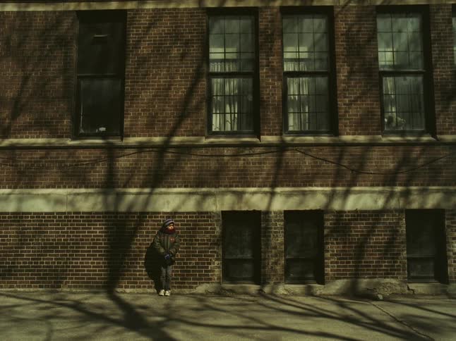 A little boy wearing a coat and hat is standing on the sidewalk by a brick apartment building