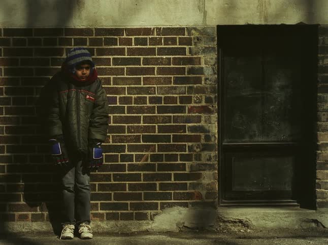 A boy bundled up in a jacket huddled against the external wall of the street feeling cold