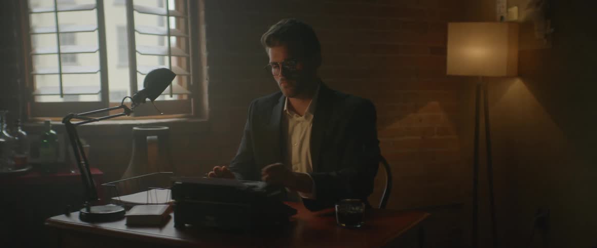 A man wearing glasses sits at a desk typing