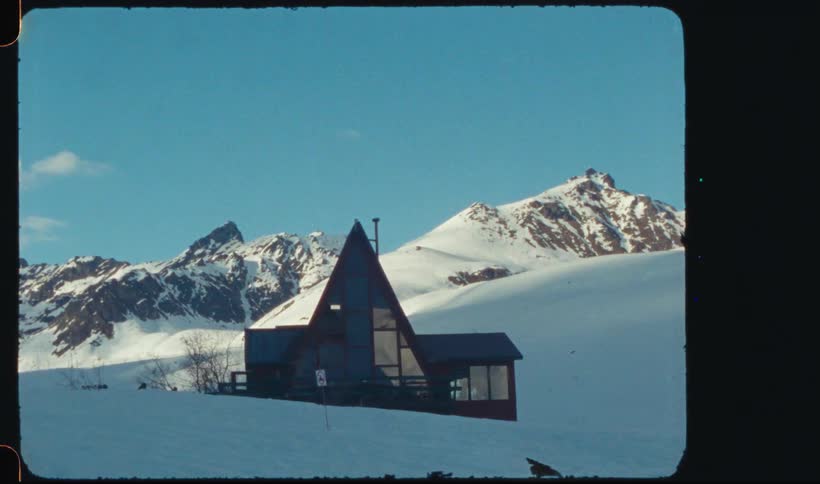Snowy scene with a house with pointed roof and mountains in the background
