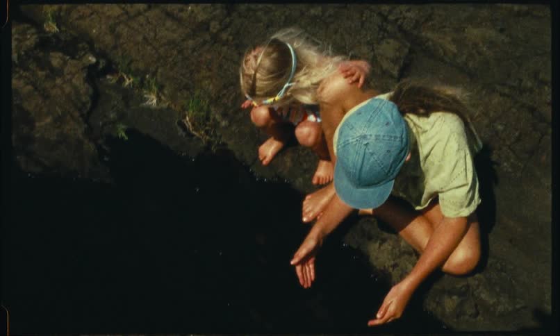Two girls crouch by a rocky shore