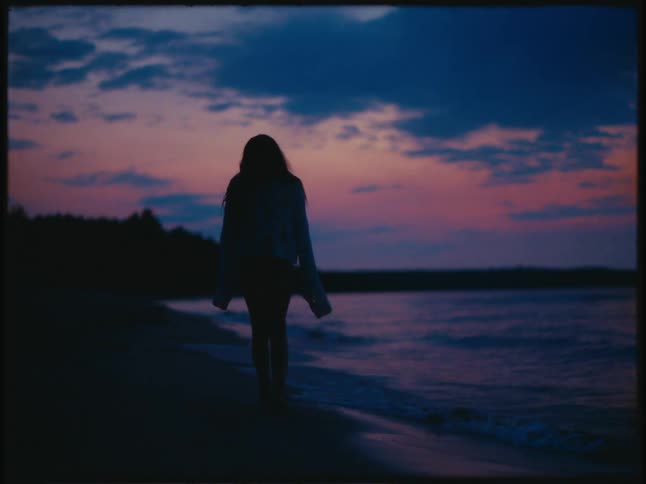 Silhouette of a woman wearing a large sweater walking on the beach at sunset