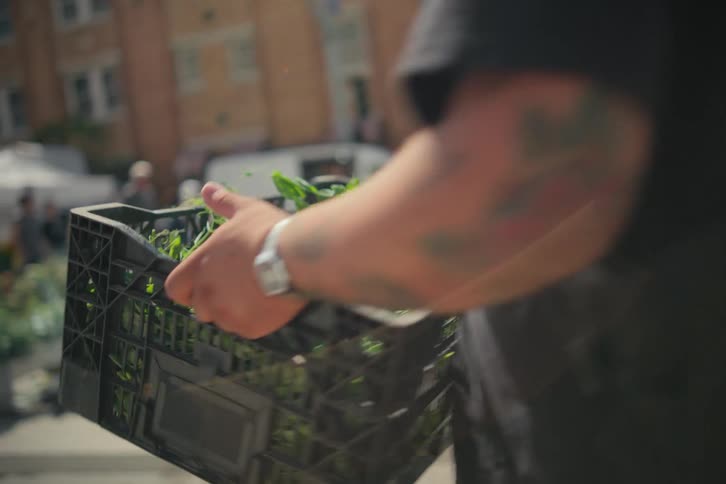 Man stacks basket of greens at outdoor market near building parking lot