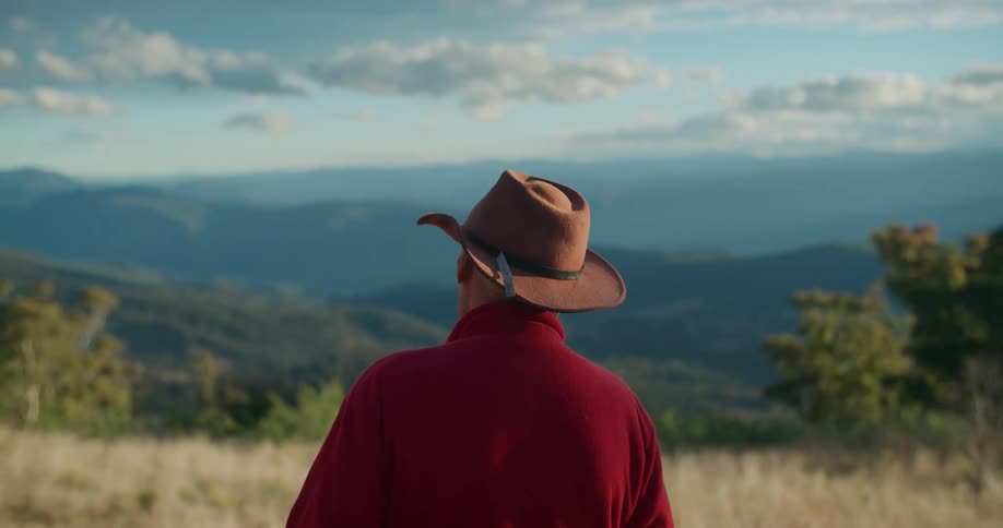 Older man wearing cowboy hat stands in field looking toward foggy mountains