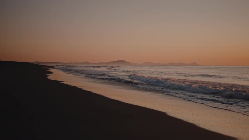 A serene and deserted beach with mountains in the distance and a yelloworange sky