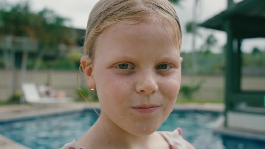 Blonde girl wet from swimming stands near swimming pool and smiles slightly