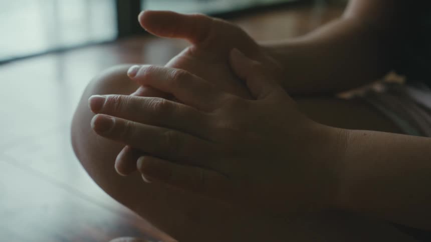 Woman indoors sitting on a wooden floor near a window touching her own hands and fingers against each other during day time