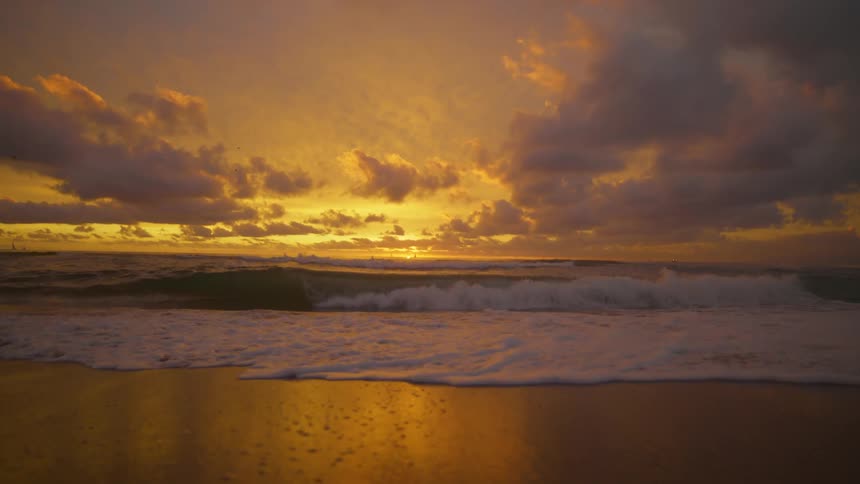 Waves splash as they roll into shore and over the beach as the sun sets on the horizon under a colorful sky