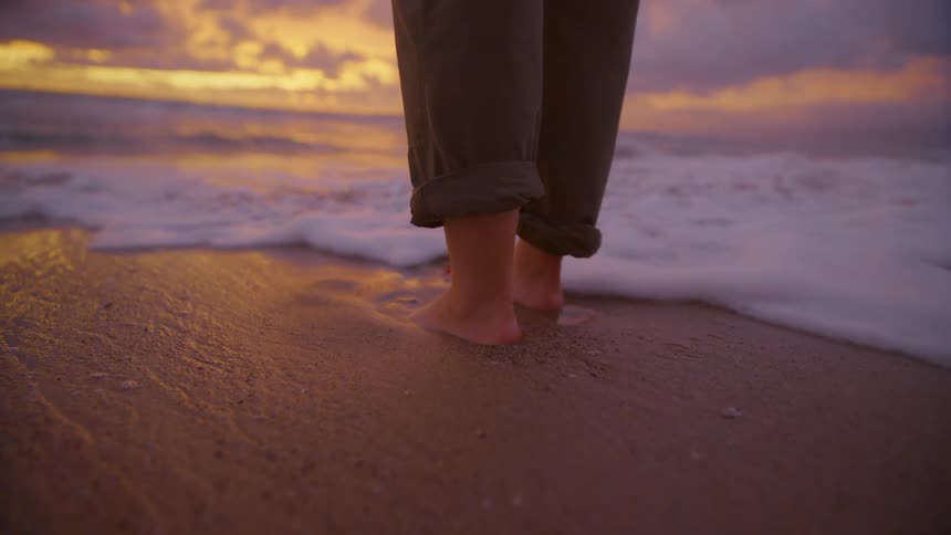 Bare feet of a person standing on the beach as waves roll into shore and over their feet as the sun sets on the horizon