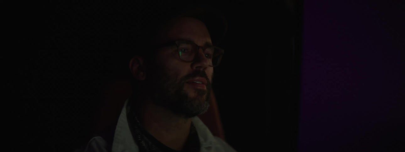 A bearded male looking at a screen in a dark room with another person