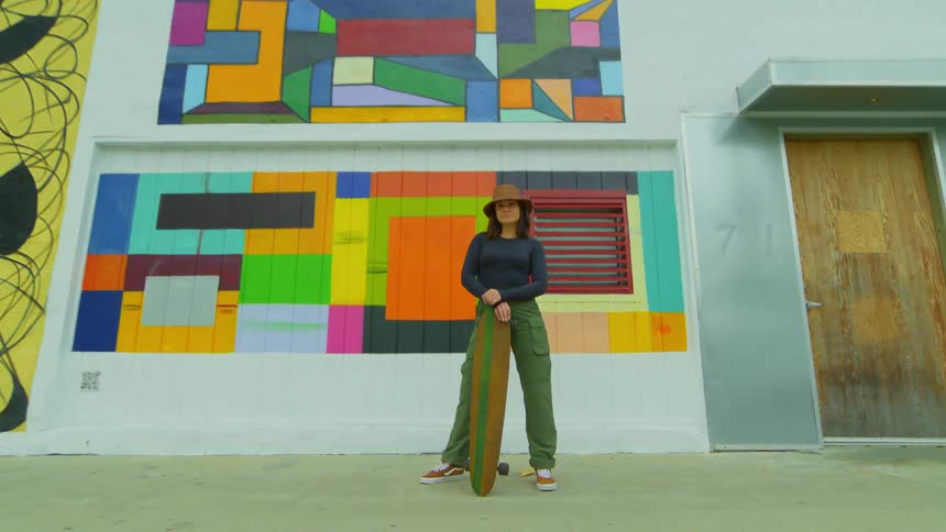 Female skateboarder stands holding skateboard in front of building decorated with colorful murals