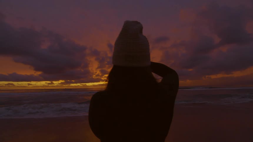 Silhouette of a woman wearing a beanie taking pictures of the sunset over the ocean