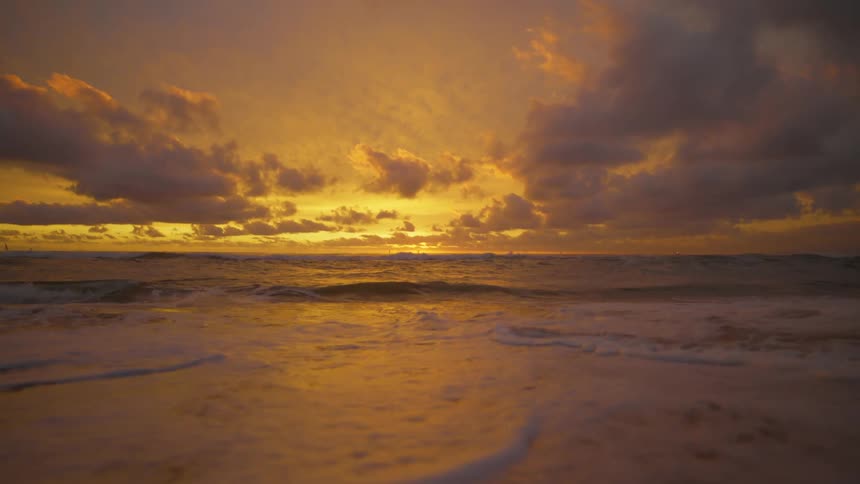 Waves splash as they roll into shore and over the beach as the sun sets on the horizon under a colorful sky