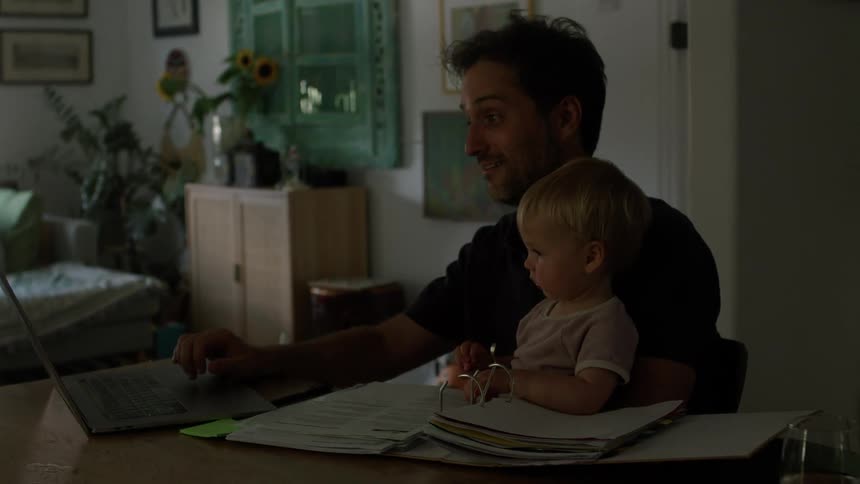 Father sitting at the table holding his baby while working on the laptop