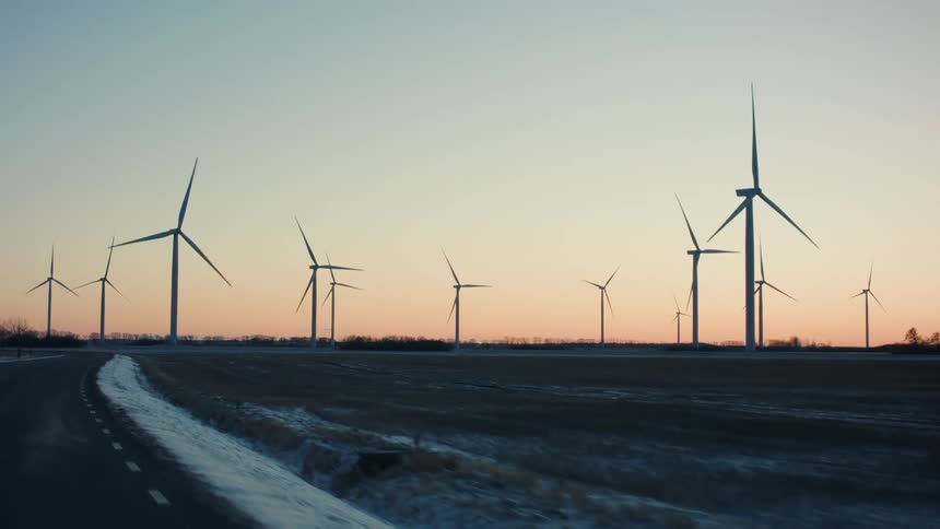 Wind turbines on icy winter field bordering highway turn slowly against a red tinted horizon